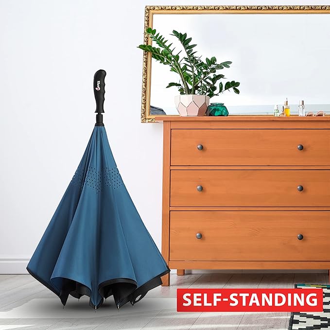 The navy blue closed Reverse Repel Umbrella standing on its own beside the wooden dresser. The umbrella is to the left and there is a ZZ Plant in a planter as well as an ornate framed gold mirror sitting atop the dresser.