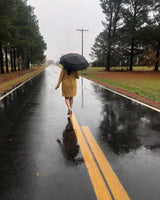 A photo of a woman holding an open umbrella walking away from the camera and down a street in the rain. 